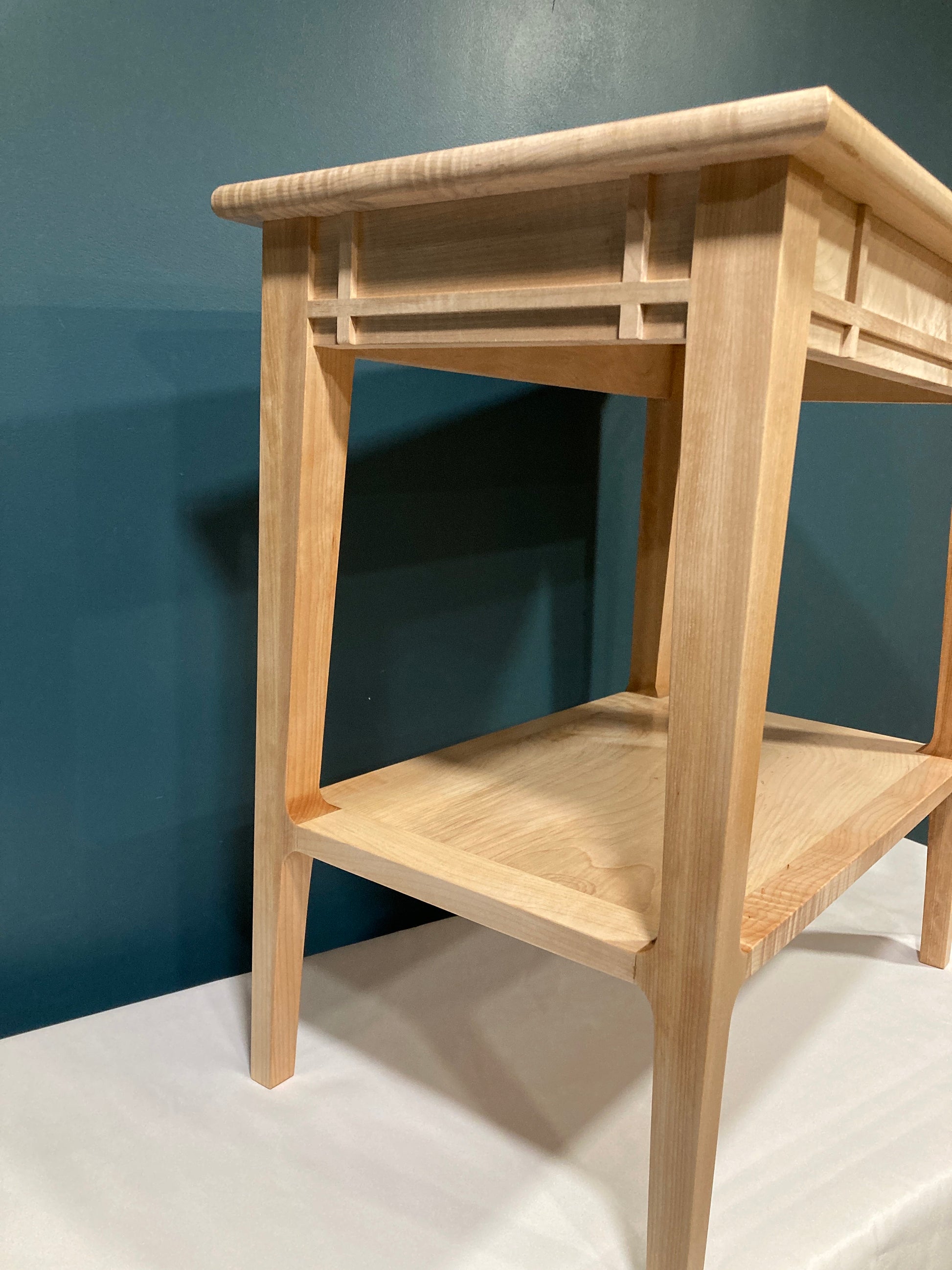  A light-colored wooden end table with a minimalist design. The table features clean lines, a simple rectangular top, and a lower shelf.  The wood grain is visible, showcasing a light, natural finish. The legs are straight and tapered slightly. The table's design is characterized by its understated elegance and functionality, suitable for a modern or contemporary interior.