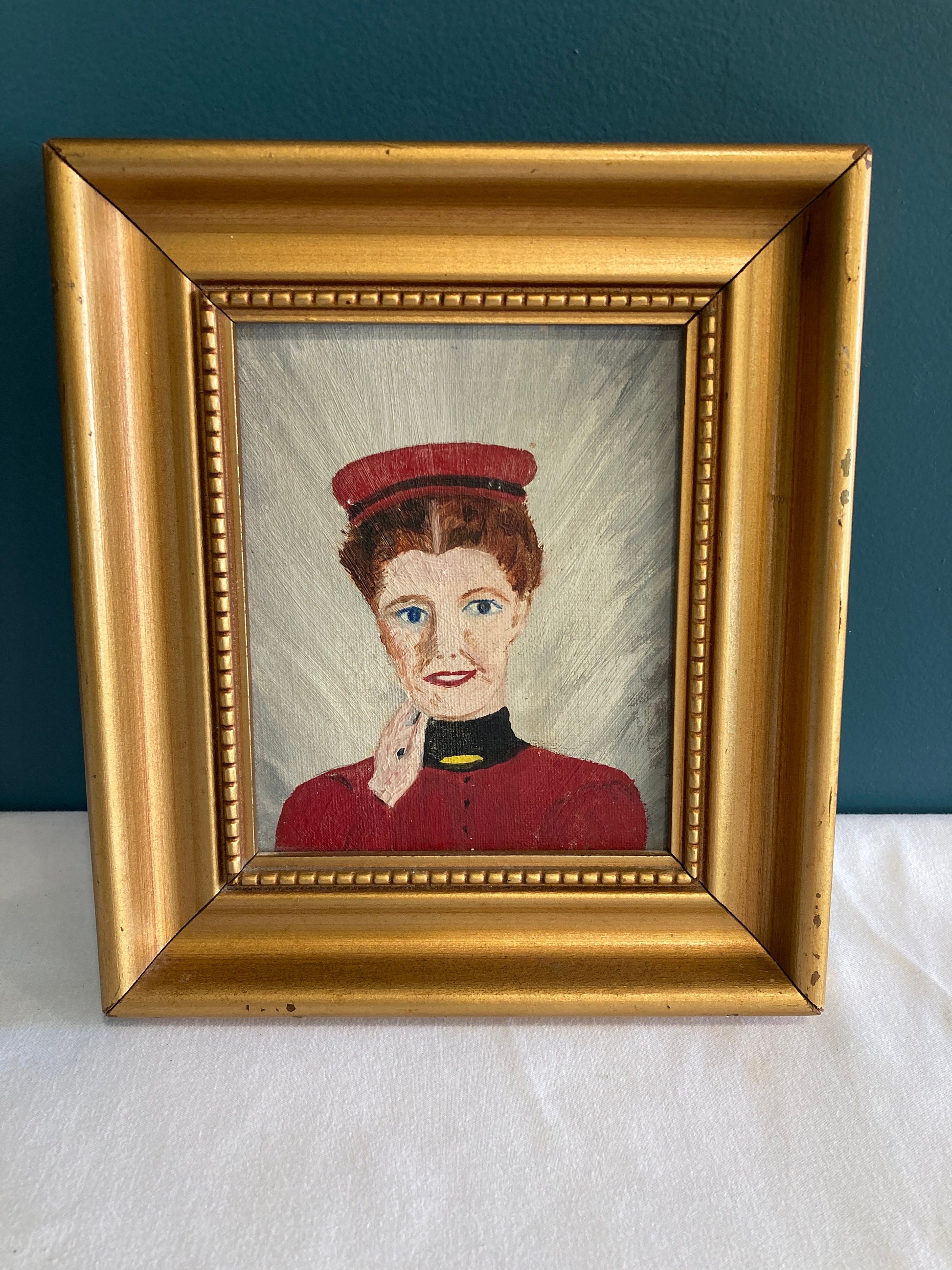 A framed painting of a woman wearing a red hat and a red dress with a black collar. The woman has her hand resting on the side of her face and is smiling and looking at the viewer.