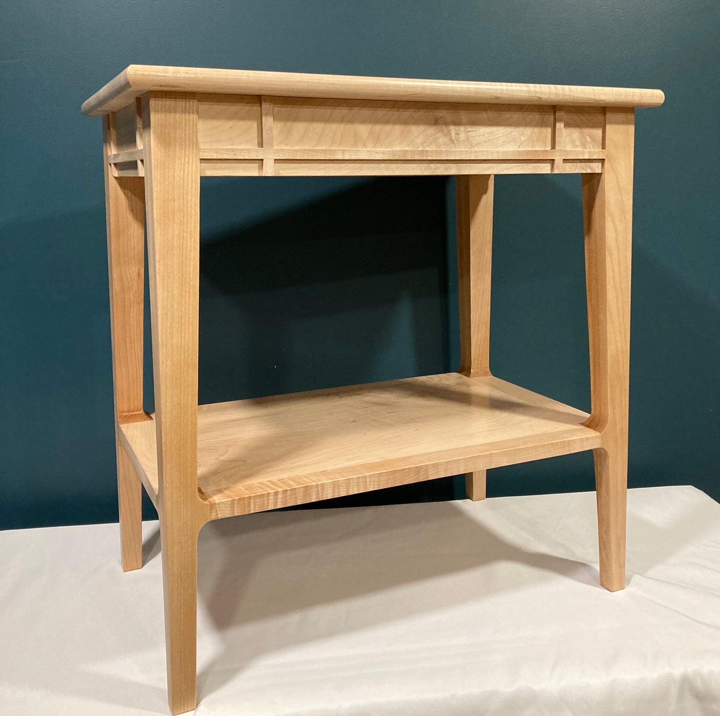 A light-colored wooden end table with a shelf. The table features a minimalist design with clean lines and a simple, rectangular shape.  The top and shelf are made of maple, showing subtle wood grain. The table's legs are straight and tapered slightly towards the bottom. The top features a decorative inset panel. This piece would be suitable for a living room or bedroom.