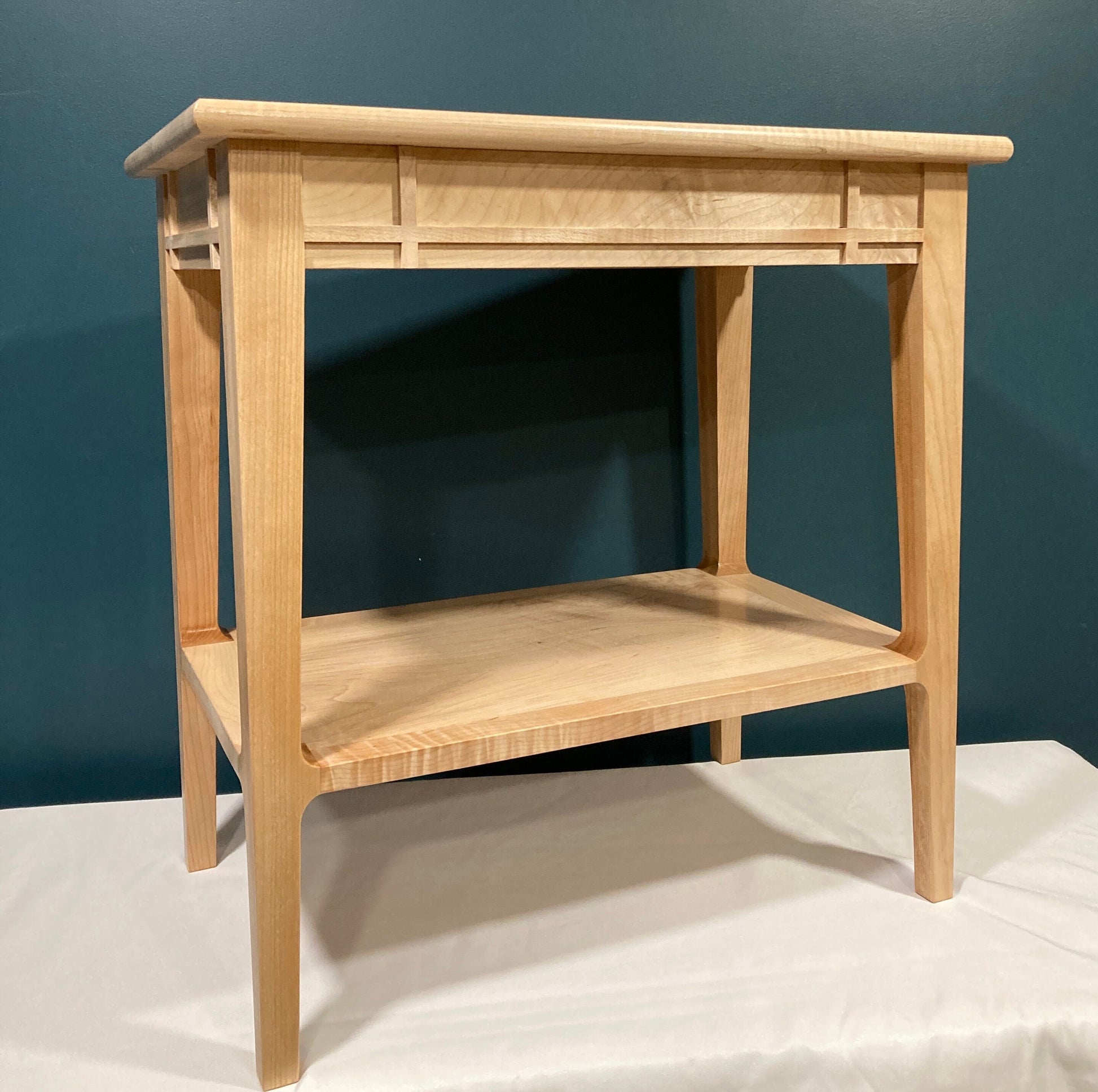 A light-colored wooden end table with a shelf. The table features a minimalist design with clean lines and a simple, rectangular shape.  The top and shelf are made of maple, showing subtle wood grain. The table's legs are straight and tapered slightly towards the bottom. The top features a decorative inset panel. This piece would be suitable for a living room or bedroom.