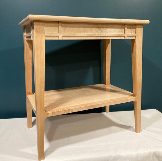 A light-colored wooden end table with a shelf. The table features a minimalist design with clean lines and a simple, rectangular shape.  The top and shelf are made of maple, showing subtle wood grain. The table's legs are straight and tapered slightly towards the bottom. The top features a decorative inset panel. This piece would be suitable for a living room or bedroom.