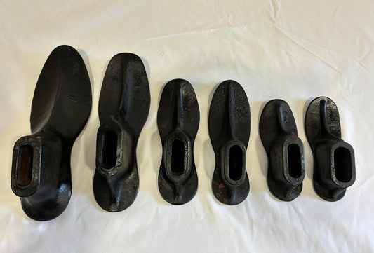 Set of six vintage cast iron shoe molds arranged in a row on white background, industrial cobbler forms with heavy black cast iron finish.