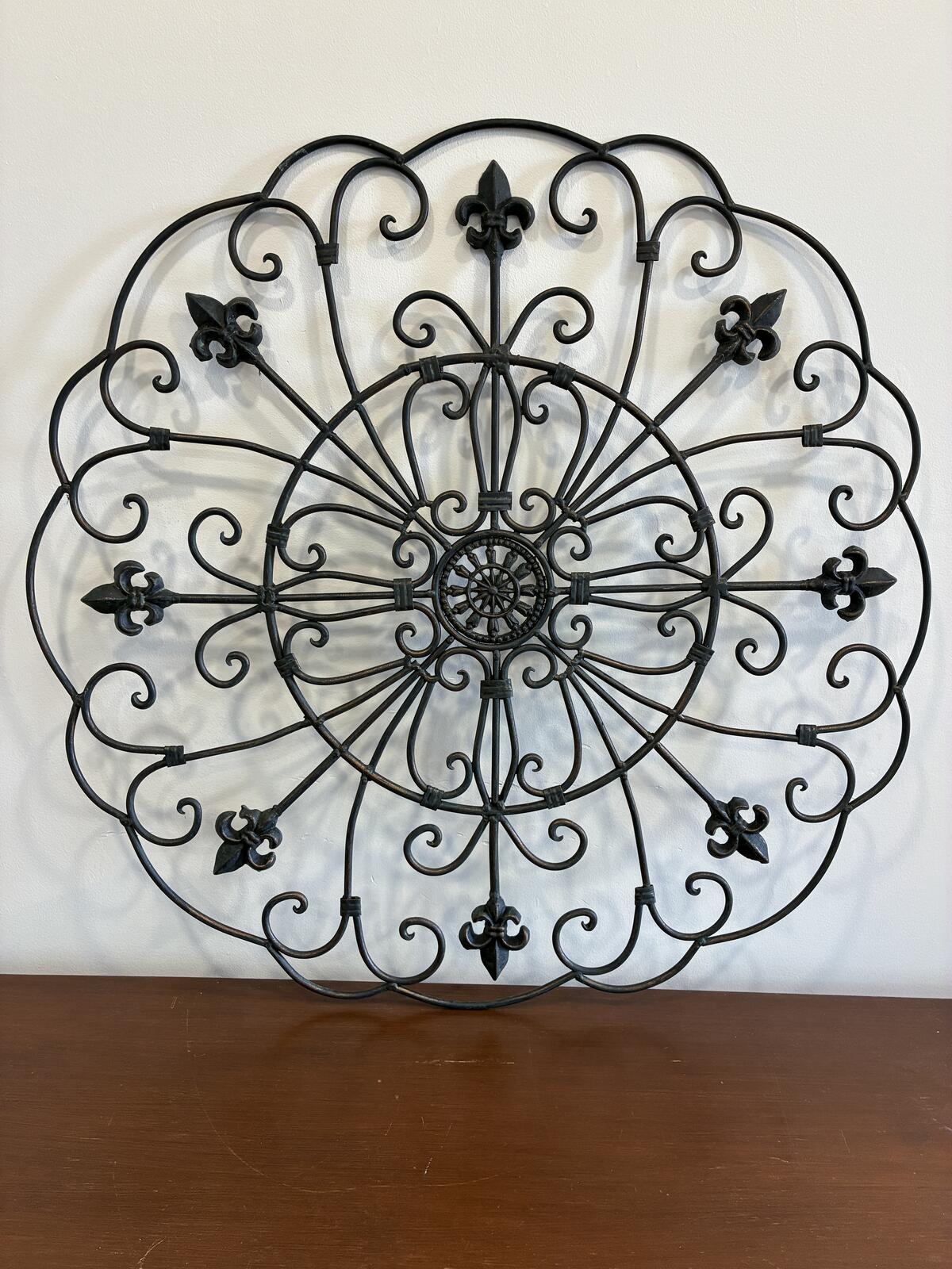  A decorative, circular wall hanging made of black wrought iron. The design features a central circular element with radiating spokes, surrounded by ornate scrollwork and fleur-de-lis accents. The piece is mounted on a white wall.