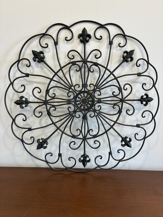  A decorative, circular wall hanging made of black wrought iron. The design features a central circular element with radiating spokes, surrounded by ornate scrollwork and fleur-de-lis accents. The piece is mounted on a white wall.