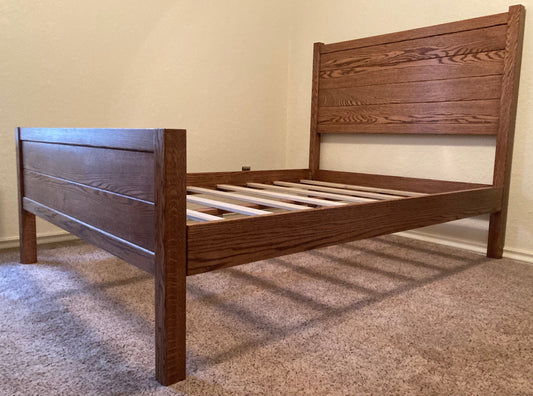 This classic style bed is Hand crafted of red oak. It has a headboard, footboard, solid wood rails, and solid wood slats.  It is sitting on beige carpet. 