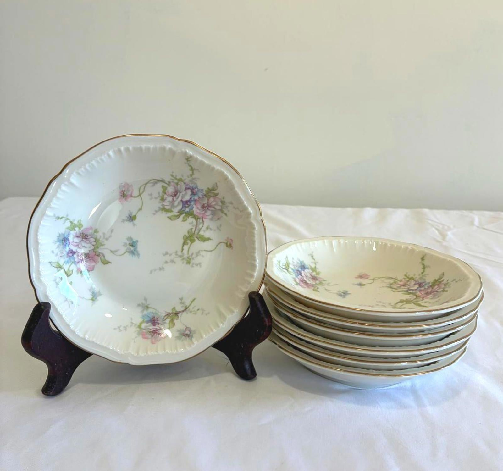 Set of seven Theodore Haviland Annette floral bowls with gold rim and scalloped edge, vintage china berry bowls. One is sitting on a display stand next to a stack of the remaining bowls.  They are on a white cloth. 