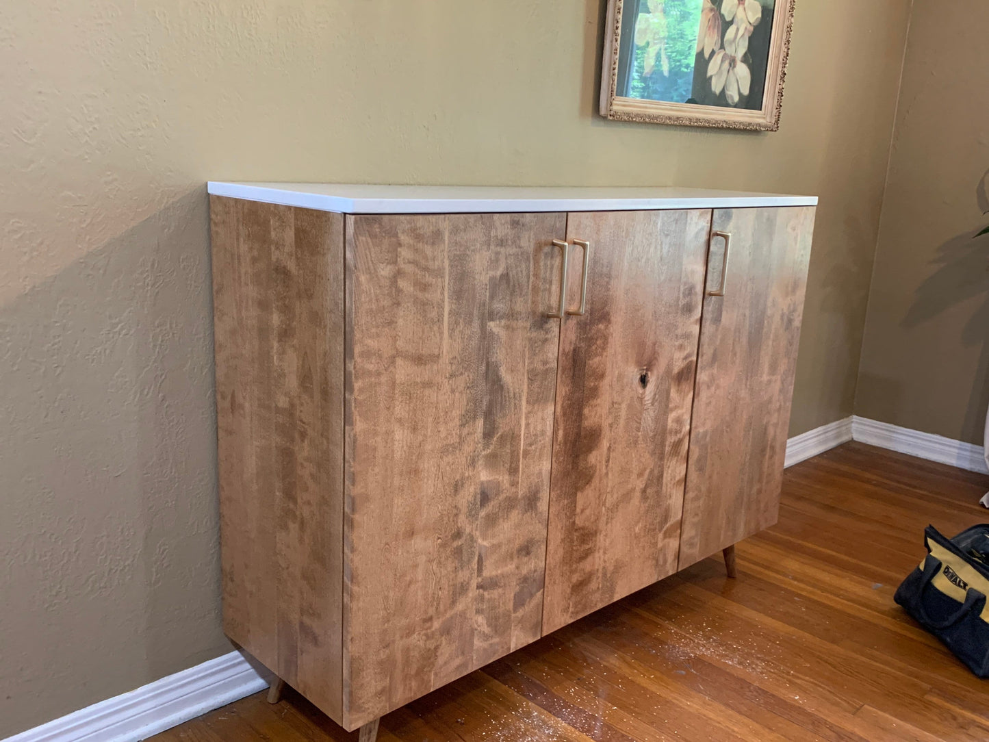 A three-door cabinet with a white quartz stone top. The cabinet is made of light brown wood with a wood grain pattern. The cabinet has gold-colored handles. The cabinet is sitting on a wooden floor.