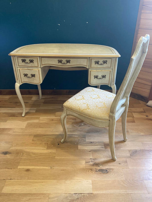 French Provincial style desk and matching chair by Sligh Furniture with leather top, cream finish, and gilt accents. The desk is sitting on a wood floor in front of a teal wall.  The matching chair has gold damask fabric on the seat.