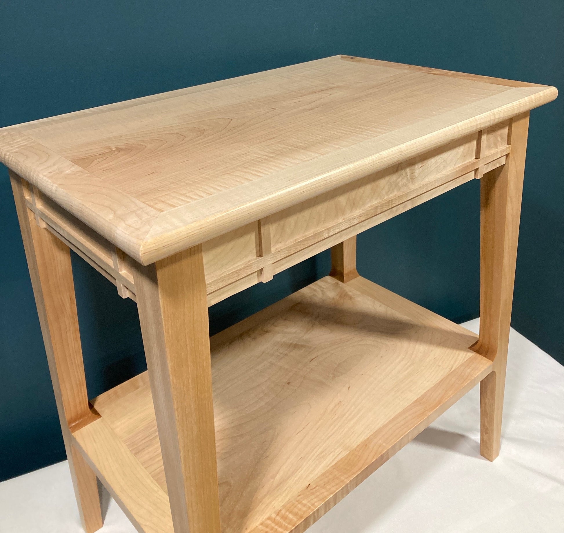 A light-colored wooden end table with a shelf. The table features a simple, clean design with a light wood finish. The top and shelf are made of wood with a subtle grain pattern. The table has a rectangular shape with straight legs and a simple frame.  Made of solid maple. The table is ideal for a living room or bedroom.