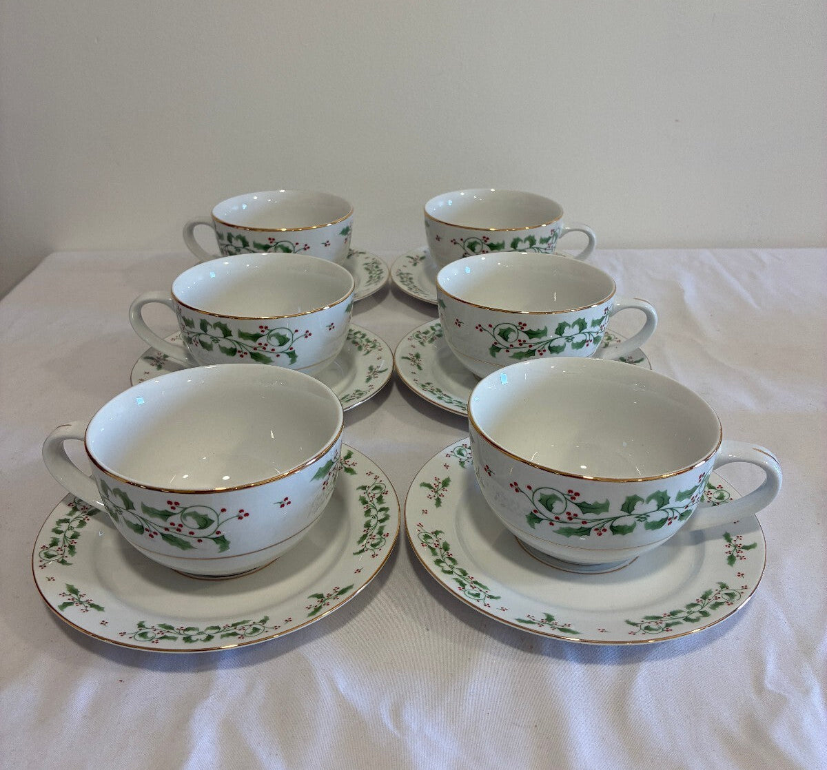 Six white porcelain teacups with matching saucers arranged on a white tablecloth. The cups and saucers are decorated with a green and red holly berry pattern and a thin gold rim.