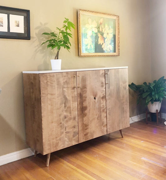  A light wood cabinet with three doors and a white quartz top. The cabinet has gold handles and sits on four wooden legs. The cabinet is in front of a light beige wall with a framed picture above it. A small green plant is on the top of the cabinet.