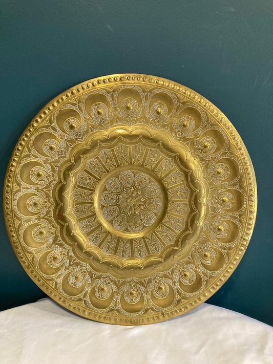 A large, round, gold-colored metal decorative plate with intricate embossed designs. The plate has a raised rim with a beaded edge. The center of the plate features a circular design with radiating lines. The plate has a shiny, metallic finish.
