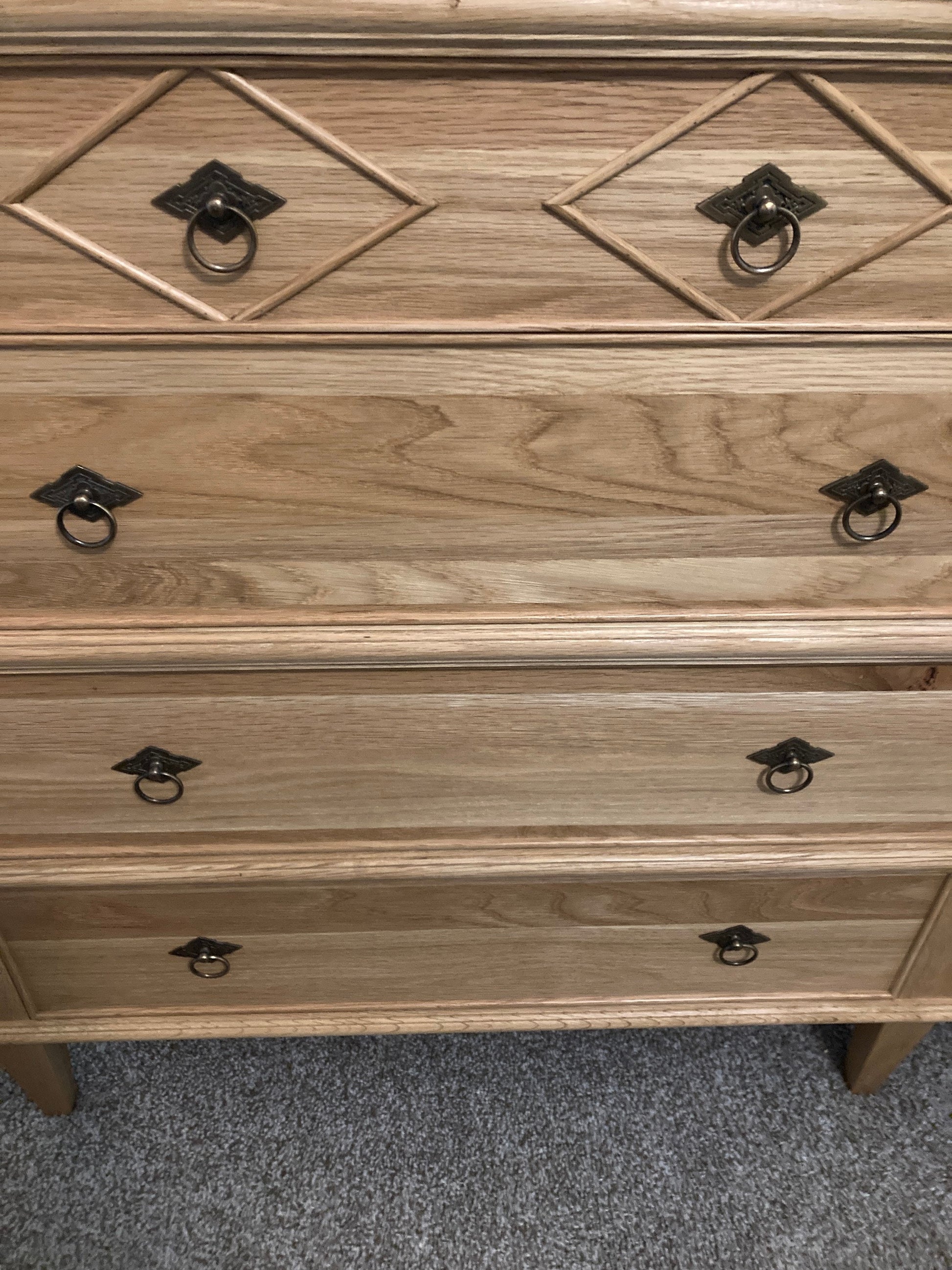 A wooden dresser with eight drawers and ornate metal ring pulls. The dresser is light brown with a natural wood finish. The dresser is sitting on a light gray carpet.