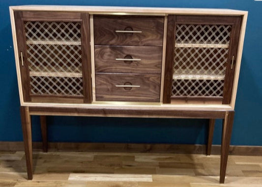Mid-century modern console cabinet handmade from mahogany and maple wood, featuring three central drawers with sleek brass pulls and two side doors with decorative brass mesh grille fronts, standing on tapered legs against a blue wall and wood floor