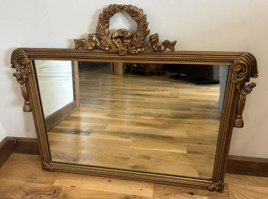 Vintage ornate gold wall mirror with carved laurel wreath crest and scrolling details leaning against a wall on hardwood floors