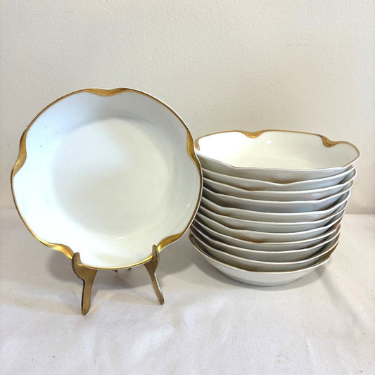 A set of white porcelain  bowls with scalloped edges and gold trim, with one bowl displayed on a small gold stand and the rest stacked on the right, against a plain off-white background.