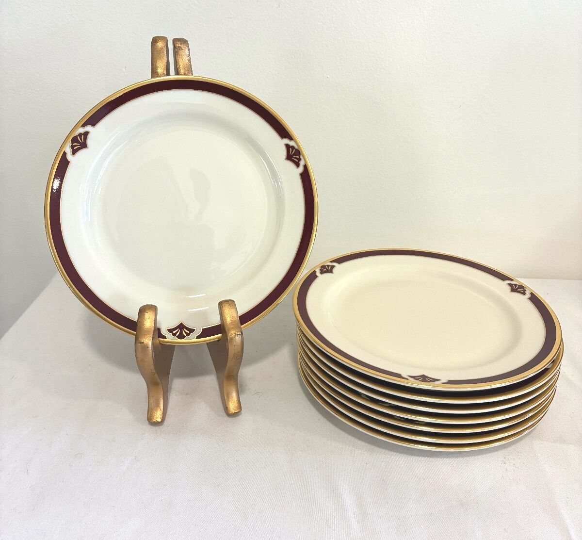 A set of white ceramic plates with gold trim and a dark red border, shown with one plate standing on a gold-colored display stand and a stack of plates beside it.