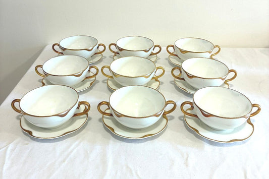A set of ten white ceramic teacups with gold trim and matching saucers, arranged in three rows of three on a white fabric surface.